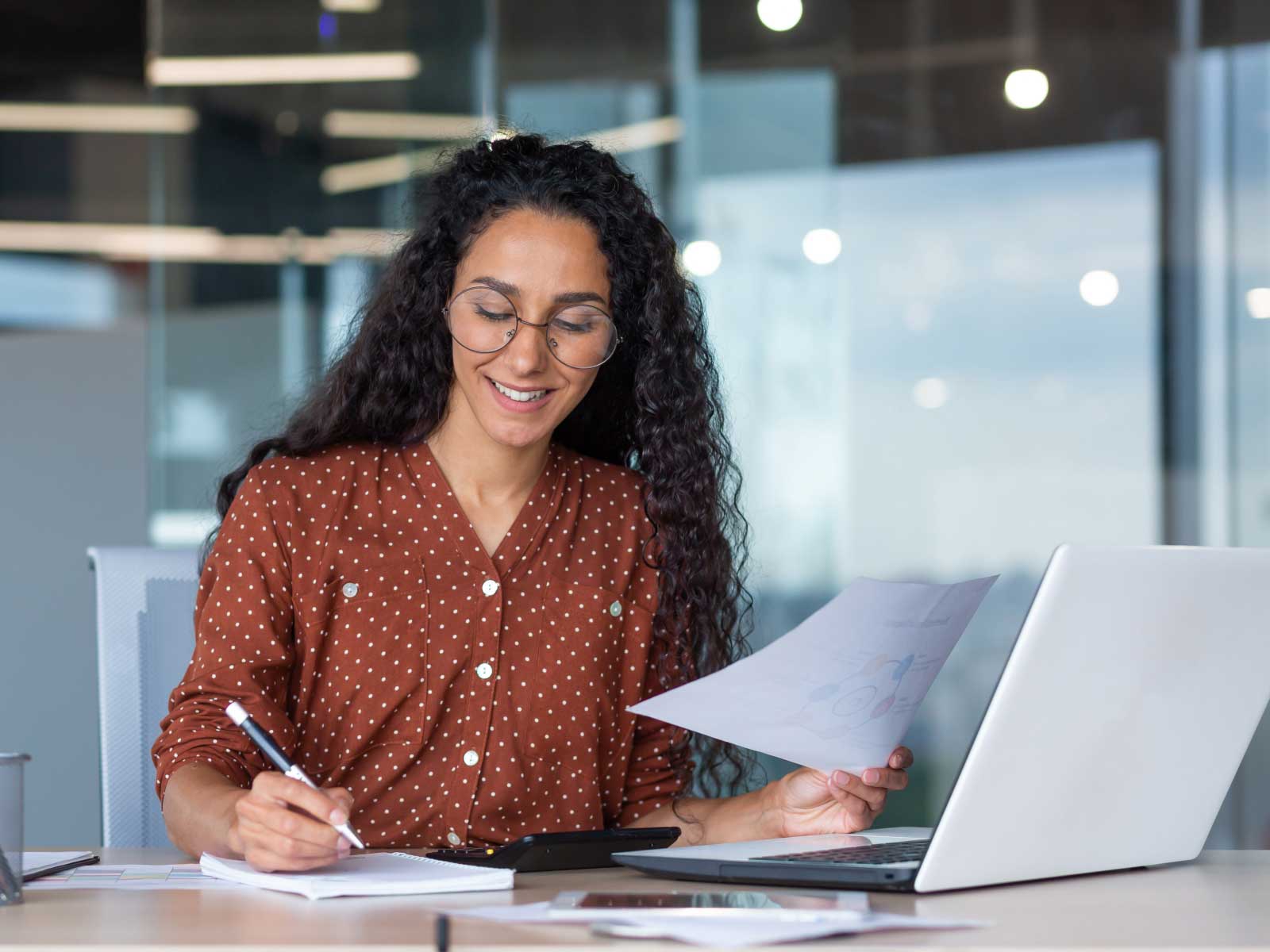 Woman writing in notebook with papers and a laptop