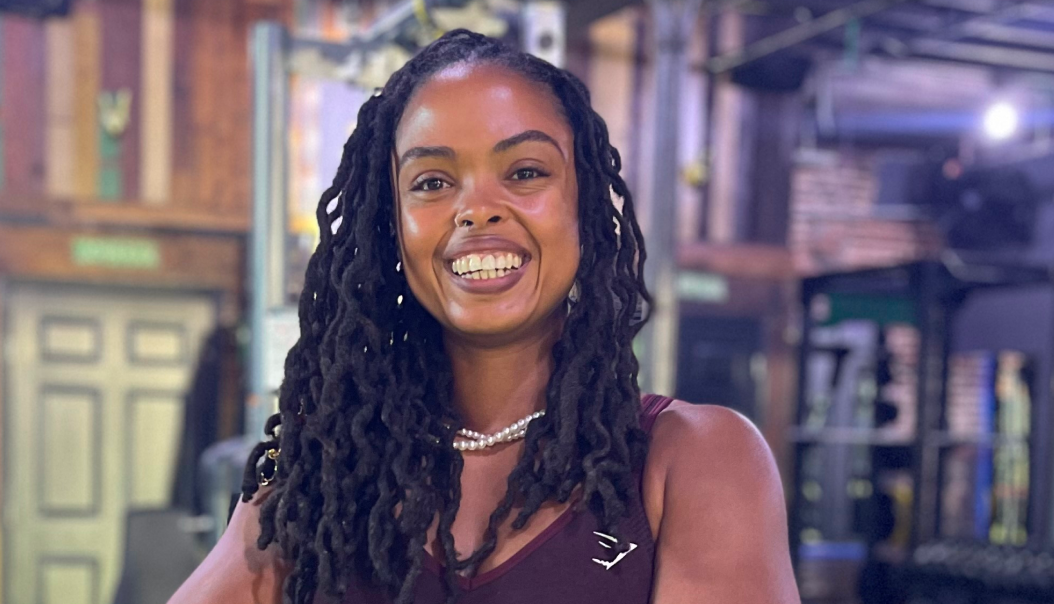 Smiling woman with long dreadlocks and a pearl necklace in a gym-like industrial studio, wearing a sleeveless top.